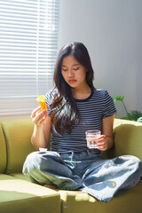 Young woman sitting on sofa at home taking pills with a glass of water, feeling sick and suffering...