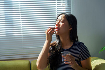 Asian woman taking pill, holding glass of water, sitting on sofa at home, sunlight through blinds