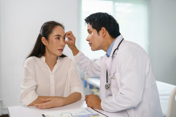 Fototapeta premium Asian doctor examining eye of female patient during medical consultation in hospital room or clinic