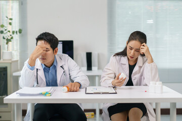 Two worried doctors are sitting at a desk, holding their heads and looking at pills, expressing...