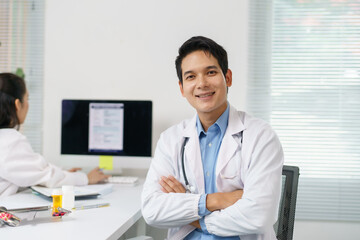 Confident doctor smiling with crossed arms, sitting in medical office, colleague working on...