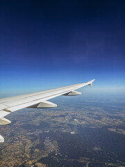 Fototapeta premium Aerial view from an airplane window showing a white aircraft wing flying over a vast rural landscape and city outskirts under a deep blue gradient sky during a daytime flight.
