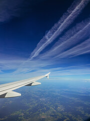 Fototapeta premium An airplane wing is visible against a vibrant blue sky with wispy contrails, soaring over a lush, green landscape, capturing the essence of air travel and adventure.