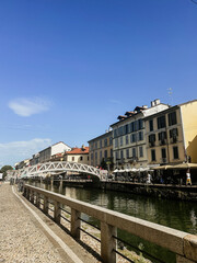 Sunny view of the Naviglio Grande canal in the vibrant Navigli district of Milan, Italy, showcasing traditional buildings, a pedestrian bridge, and the cobblestone walkway along the water.