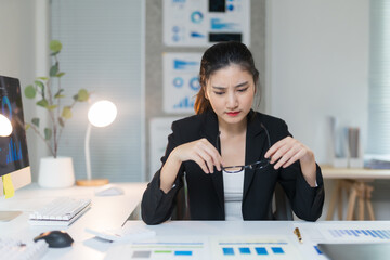 Young Asian manager analyzing accounting paperwork, facing challenges from declining sales while feeling stressed and overworked at her desk