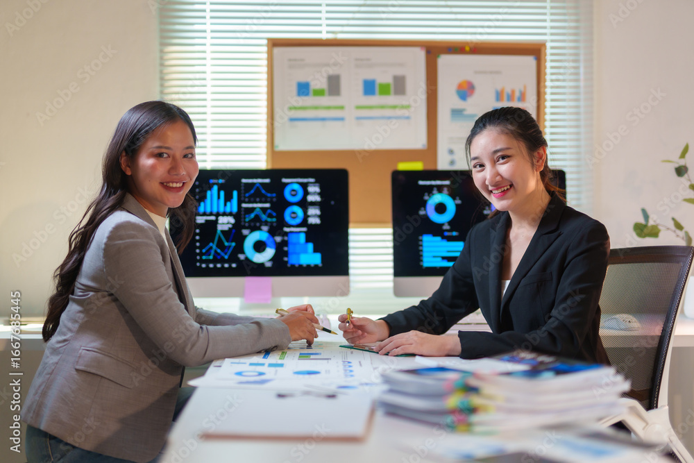 Poster two smiling businesswomen working together analyzing financial data using computer and printed repor