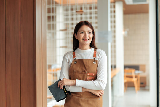 Portrait of asian waitress standing with arms crossed and holding napkin, looking away and smiling in coffee shop - Powered by Adobe