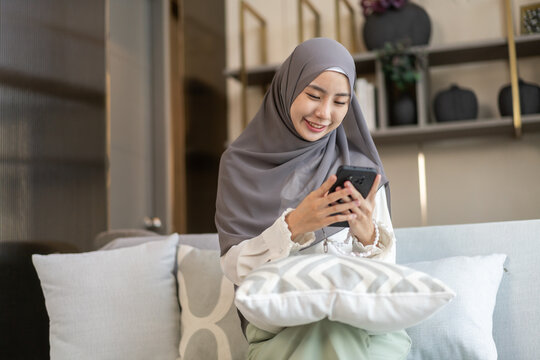 Asian muslim woman use smartphone at home, female in hijab checking messages, modern communication, smiling and holding mobile device technology use, staying connected, lifestyle concept, social media - Powered by Adobe