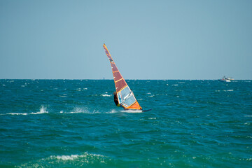 A windsurfer with a transparent orange sail glides across deep blue-green waves under a clear sky,...