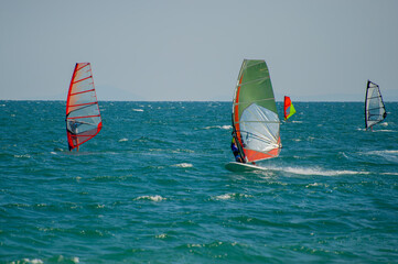 A group of windsurfers with colorful sails glide across choppy blue-green waters under a clear sky, framed by distant islands on the horizon in Bulgaria