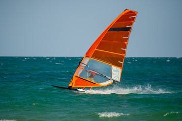 A windsurfer glides across the ocean with a vibrant orange sail under a clear sky in Bulgaria