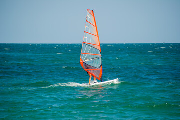 A windsurfer with a transparent orange sail glides across deep blue-green waves under a clear sky,...