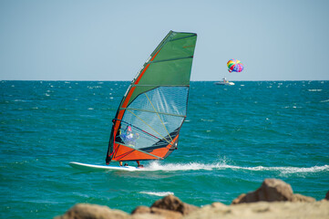 A windsurfer skims across turquoise waves with a vibrant orange and green sail, while a parasailer...