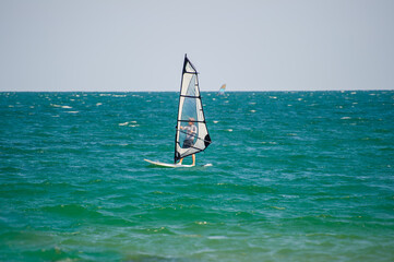 A windsurfer with a transparent sail glides over turquoise waves under a clear sky, with another...