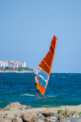 A windsurfer with a vivid orange sail glides through ocean waves near a rocky coast and beachfront...