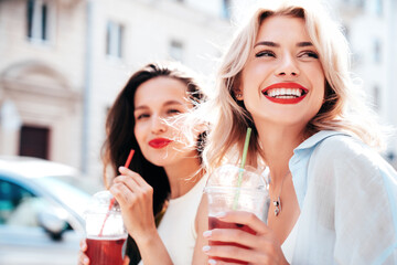 Two young beautiful smiling glam female in trendy summer clothes. Sexy women posing outdoors. Positive models holding and drinking fresh cocktail smoothie drink in plastic cup. Bright makeup, red lips