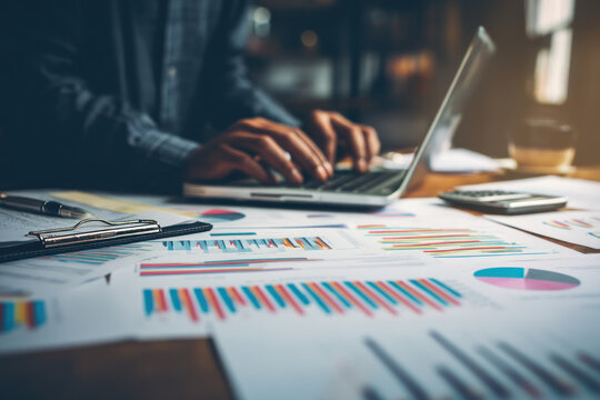 A fintech entrepreneur reviewing graphs on a laptop. Stacks of reports and financial data cover the table