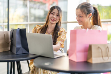 Two diverse women smiling and talking while use laptop for online shopping at cafe, searching for deals and discounts with shopping bags on table, concept of e-commerce, retail, digital lifestyle