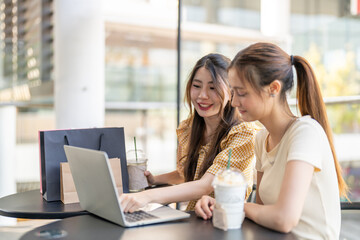 Two diverse women smiling and talking while use laptop for online shopping at cafe, searching for deals and discounts with shopping bags on table, concept of e-commerce, retail, digital lifestyle