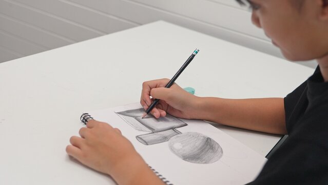 Student practicing pencil drawing of 3D geometric shapes in sketchbook