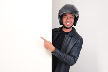 A motorcycle rider wearing helmet peeking behind wall with happy face and pointing beside him