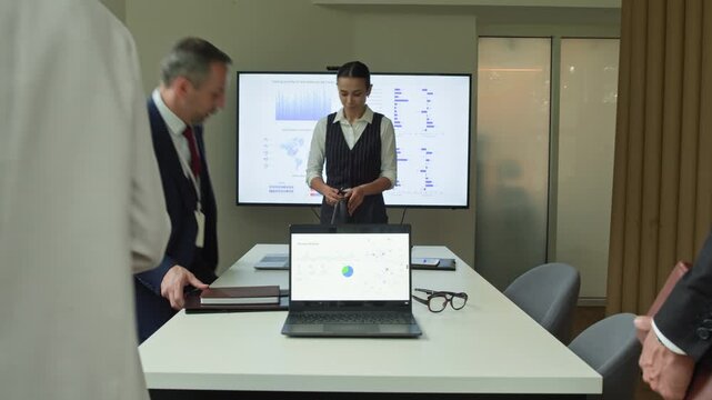 Young female business coach entering boardroom or office with interactive board while group of diverse employees in formalwear walking up to table and taking their seats before seminar