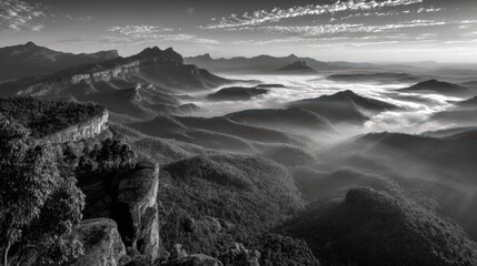 Sunrise over rugged mountain landscape with misty valleys and cloud-covered peaks in black and white