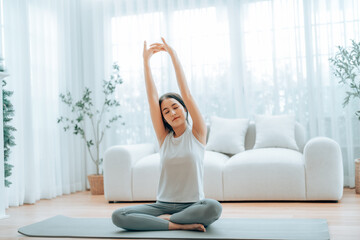Happy young Asian woman practicing yoga and shoulder stretching at home sitting on floor in living room
