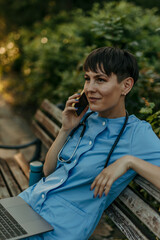 Female doctor relaxing on bench using laptop and talking on phone
