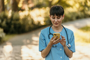 Female doctor using smartphone in park wearing blue scrubs and stethoscope
