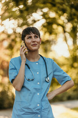 Female doctor wearing blue scrubs is talking on phone in a park
