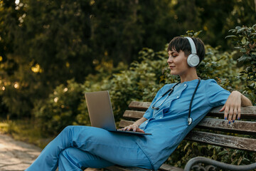 Doctor wearing headphones and using laptop while relaxing on bench in park