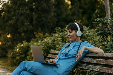 Doctor wearing headphones and using laptop while relaxing on bench in park