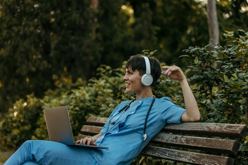 Doctor relaxing on bench with laptop and headphones in park