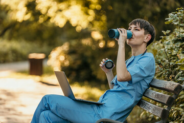 Doctor drinking coffee and relaxing on bench in park after work with laptop