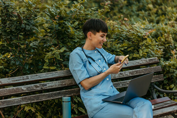 Smiling doctor using smartphone and laptop on bench in park