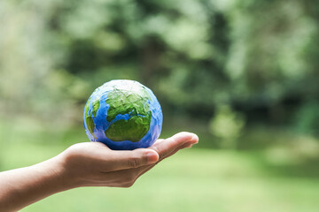 Hand holding a painted paper model of Earth with green natural background, symbolizing...
