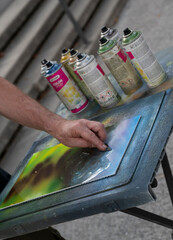 A talented artist applies spray paint to a canvas, showcasing a colorful landscape. Nearby, spray cans in various colors hint at the artwork's dynamic process, set against a concrete backdrop.