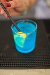 A refreshing blue cocktail with a lemon slice and a black straw being stirred in a glass on a bar counter
