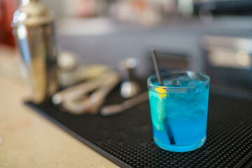 Vibrant Blue Cocktail Garnished with Lemon Slice and Black Straw on a Bar Counter with Shaker and Tools in Background Ideal for Refreshment and Celebration