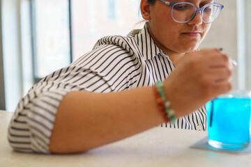 Close Up Of A Woman Wearing Glasses Stirring A Bright Blue Cocktail In A Glass With A Spoon With A...