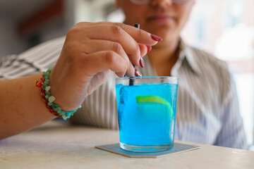 Cheerful woman mixing bright blue cocktail, adorned with colorful bracelets, relaxing at bar with...