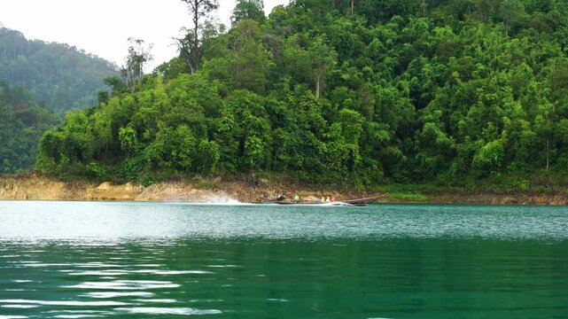A scenic wide shot of a traditional long-tail boat gliding across a serene lake, surrounded by towering, lush green mountains and dense tropical rainforest. The boat creates gentle ripples in the calm