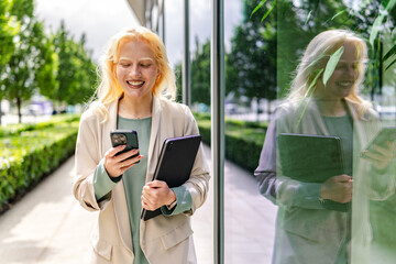 Albino woman smiles while using smartphone and holding a tablet near a glass building on a sunny day