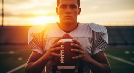Young male american football player holding ball on stadium field at sunset with strong determined expression. Sportsman portrait for athletic competition.