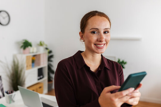 Businesswoman smiling while using smartphone indoors at the office
