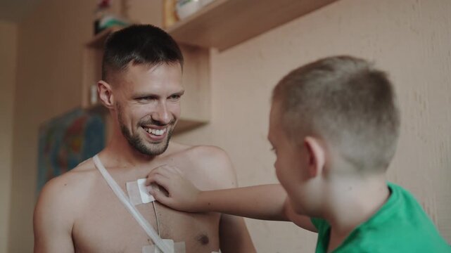 Caucasian boy touches Holter monitors on smiling man chest with hands. Son asks about father health and methods of diagnosing heart abnormalities at home. Health care and family support.