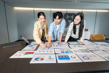 Group of asian coworkers standing around a table during a team brainstorming session, reviewing ideas and charts together to support project success through collaborative team brainstorming