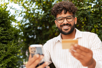 Smiling young man making online payment using credit card and smartphone outdoors