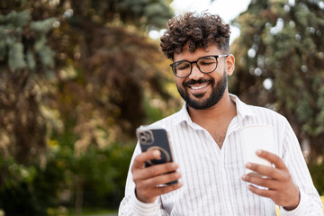 Fototapeta premium Young Indian Man Using Smartphone and Holding Coffee in Park